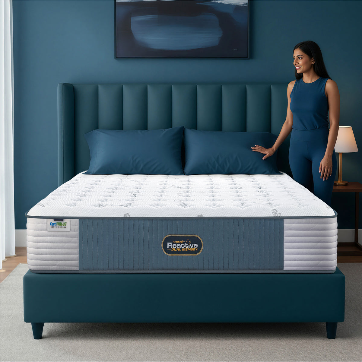 Springfit Reactive Dual Memory mattress lifestyle image with woman in teal outfit standing beside mattress on teal upholstered channeled headboard bed in deep blue accent bedroom with side lamp